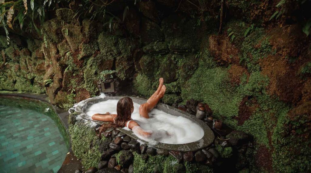 a tourist relaxing in a bubble-filled stone bathtub surrounded by a mossy tropical jungle wall in Ubud, Bali.